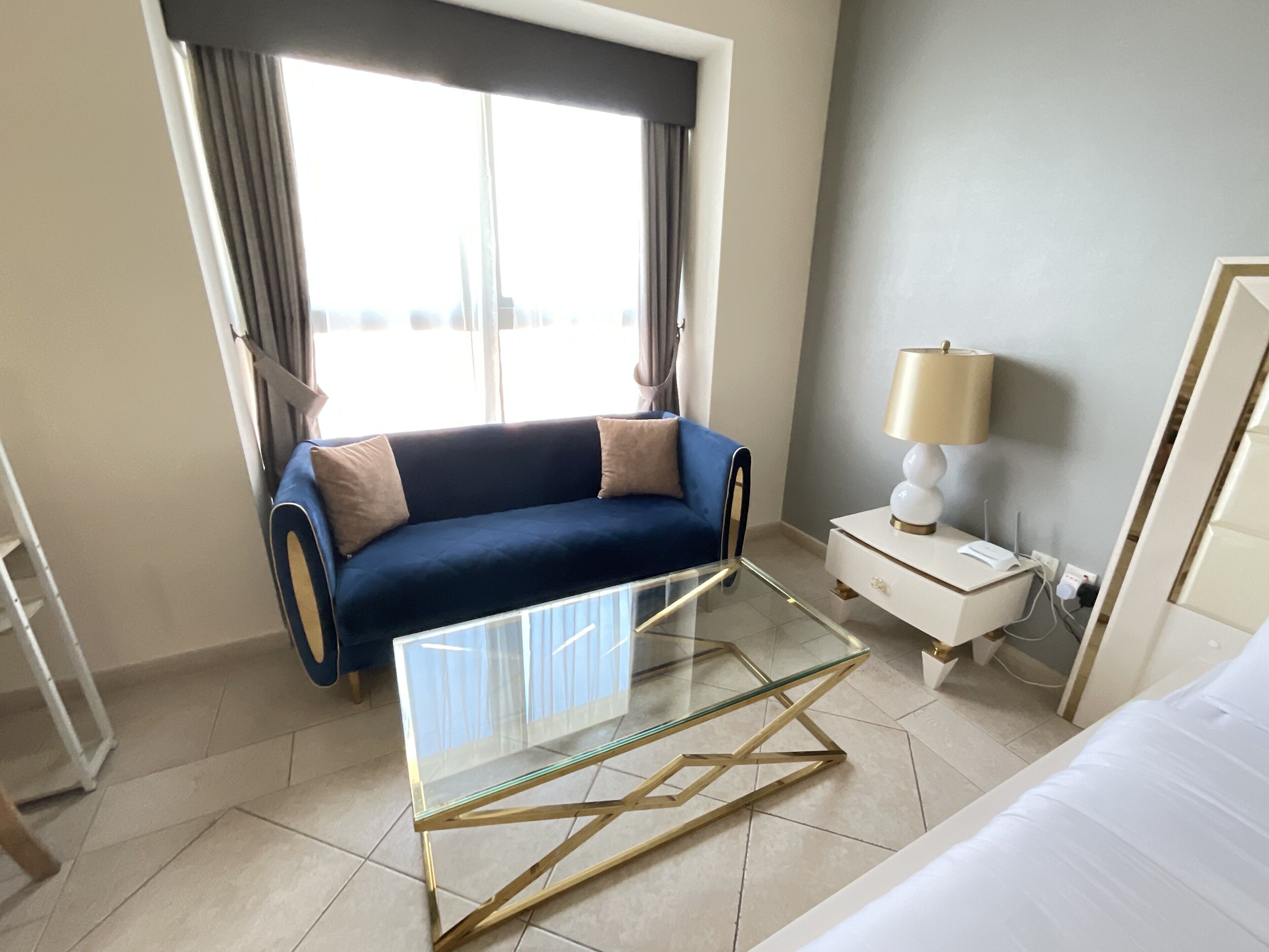 Une chambre d’hôtel moderne avec un canapé bleu, une table basse en verre, une table de chevet blanche avec une lampe, et une vue sur la ville grâce à de grandes fenêtres.