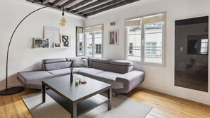 A modern living room with a grey sectional sofa, a dark coffee table, and a floor lamp. There are framed pictures on the walls and large windows providing natural light. - Chic 2BR by Rue Montorgueil - Les Halles (Paris)