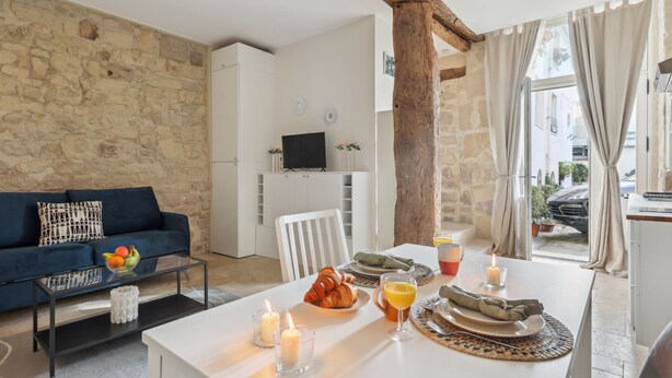 A modern living room with a stone wall, a dark blue sofa, a glass coffee table, and a dining area with a white table set for breakfast. - Cosy Studio in the Heart of Ile Saint Louis (Paris)