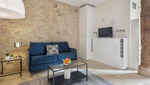 A modern living room with a stone wall, a blue sofa, a glass coffee table with fruit, a TV, and a white cabinet. - Cosy Studio in the Heart of Ile Saint Louis (Paris)