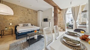 A bedroom with a stone wall, a bed, a bedside table with a lamp, and a TV. There is a dining area with a table set for two, plates, and a glass of juice. - Cosy Studio in the Heart of Ile Saint Louis (Paris)