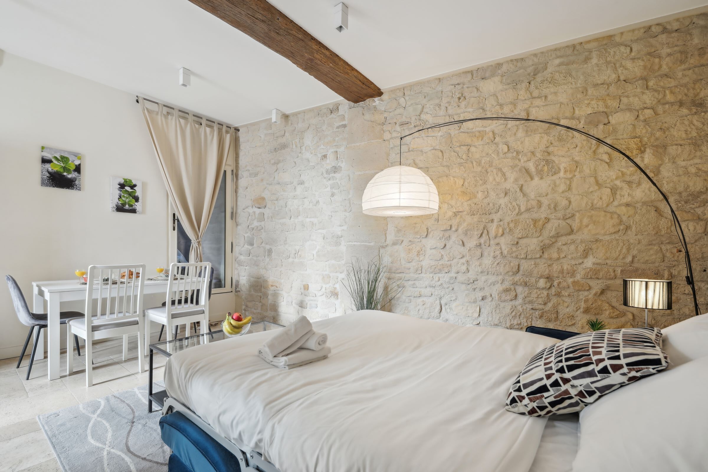 Een slaapkamer met een stenen muur, een bed met wit beddengoed, een bureau met stoelen en een hangende lamp.