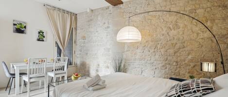 Een slaapkamer met een stenen muur, een bed met wit beddengoed, een bureau met stoelen en een hangende lamp.