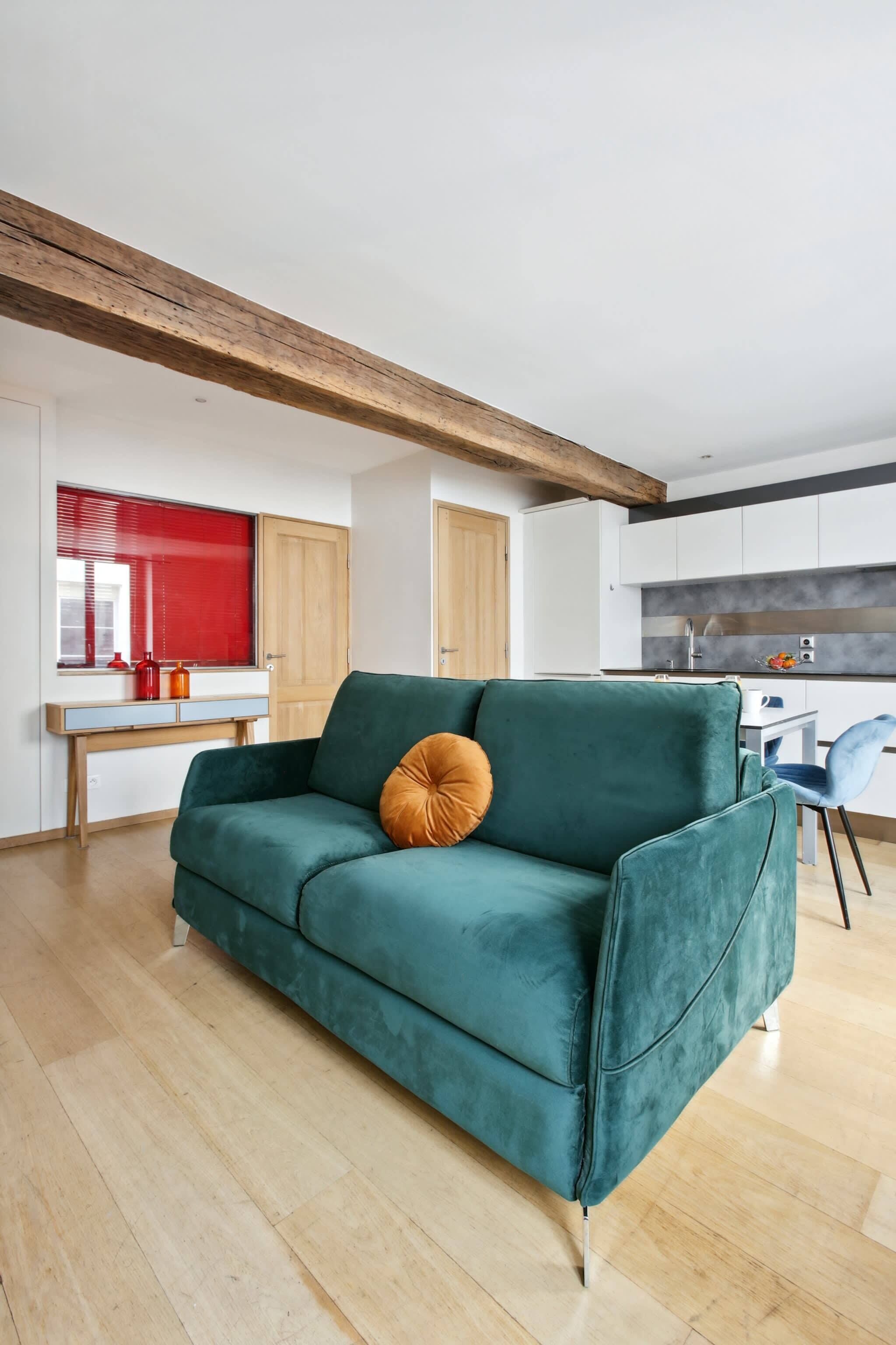Un soggiorno moderno con un divano verde acqua, una trave in legno a soffitto, una persiana rossa e una cucina sullo sfondo.