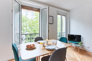 A dining area with a table set for breakfast, a TV, and a balcony with a view of greenery. - Spacious 1BR next to Montmartre (Paris)