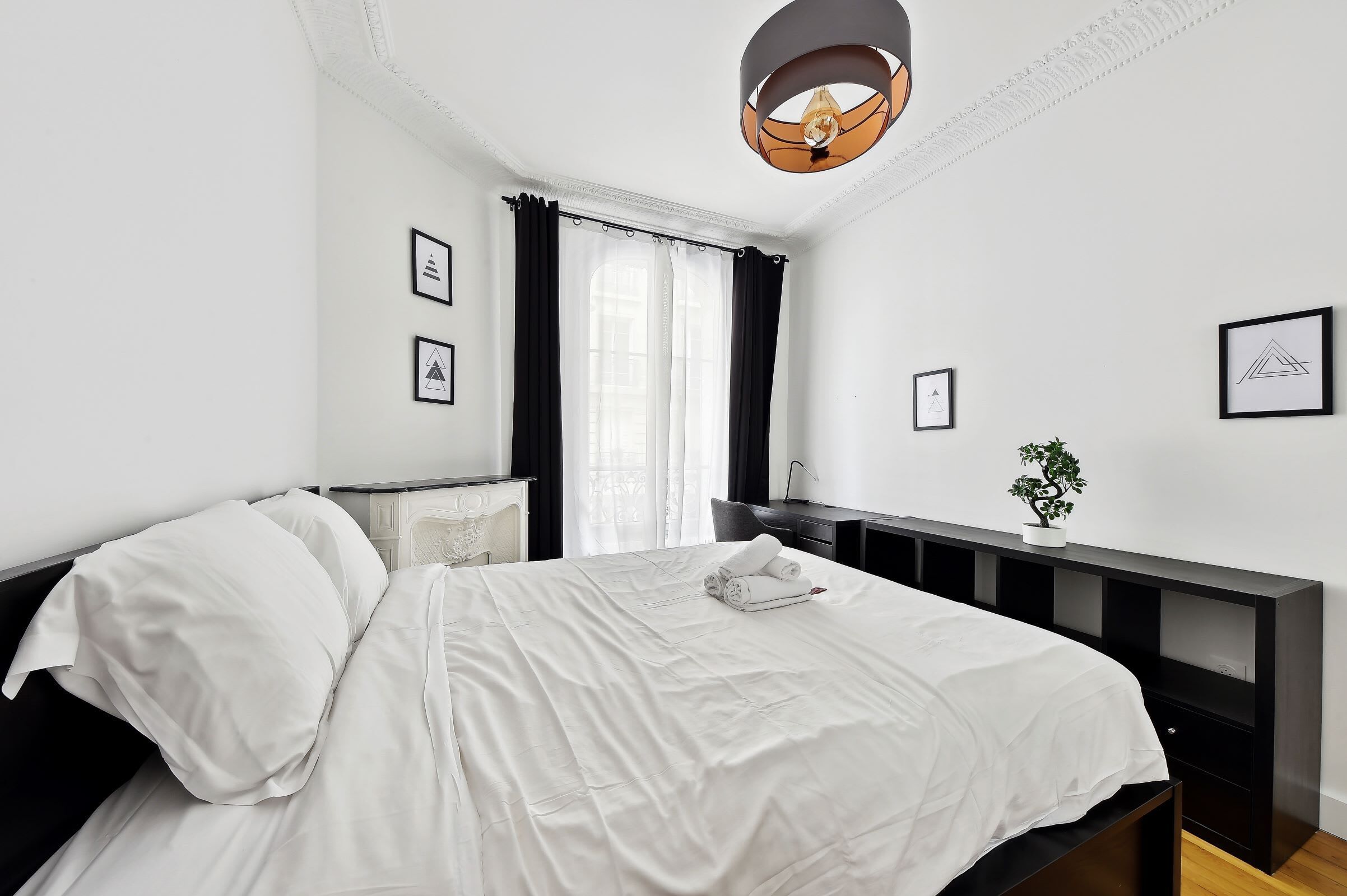 Una cama bien hecha con ropa blanca, una mesita de noche con una lámpara y una planta en maceta sobre una consola negra.