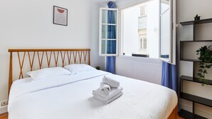A neatly made bed with white linens and a folded towel, a wooden headboard, a window with blue curtains, and a small plant on a shelf. - 1BR with Balcony - Latin Quarter & Blvd St Germain (Paris)