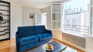 A modern living room with a blue sofa, a glass coffee table with fruit, and a large window with a city view. - 1BR with Balcony - Latin Quarter & Blvd St Germain (Paris)
