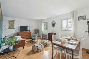 A modern living room with a sofa, ottomans, a coffee table, and a TV. There is a dining area with a table set for two, and a window with curtains. - Spacious 1BR In the Latin Quarter - Mouffetard (Paris)