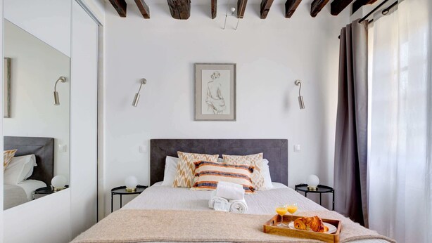 A neatly made bed with a tray of croissants and orange juice, a framed picture on the wall, and a mirror on the left.
