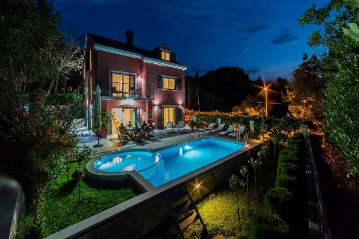 A two-story house with a swimming pool, surrounded by greenery and illuminated by night lights.