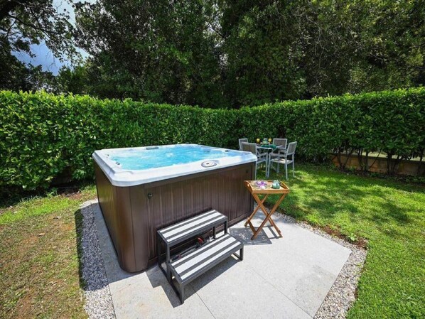 A hot tub with a bench and a table with drinks, surrounded by a hedge and grass.