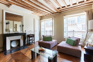 A modern living room with a glass coffee table, two sofas, a fireplace, and large windows with a view of buildings.