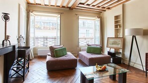 A living room with wooden flooring, two sofas with green cushions, a glass coffee table with fruits, a lamp, and a view of buildings through large windows. - Spacious 2BR between Place Vendome and Madeleine (Paris)