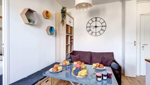 A dining area with a glass table set with croissants, fruits, and beverages. There is a clock on the wall and a bookshelf with plants.