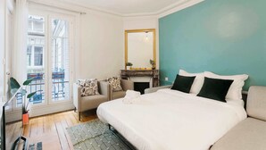 Une chambre à coucher comprenant un lit, un canapé, une petite table, un miroir et une fenêtre donnant sur des immeubles.