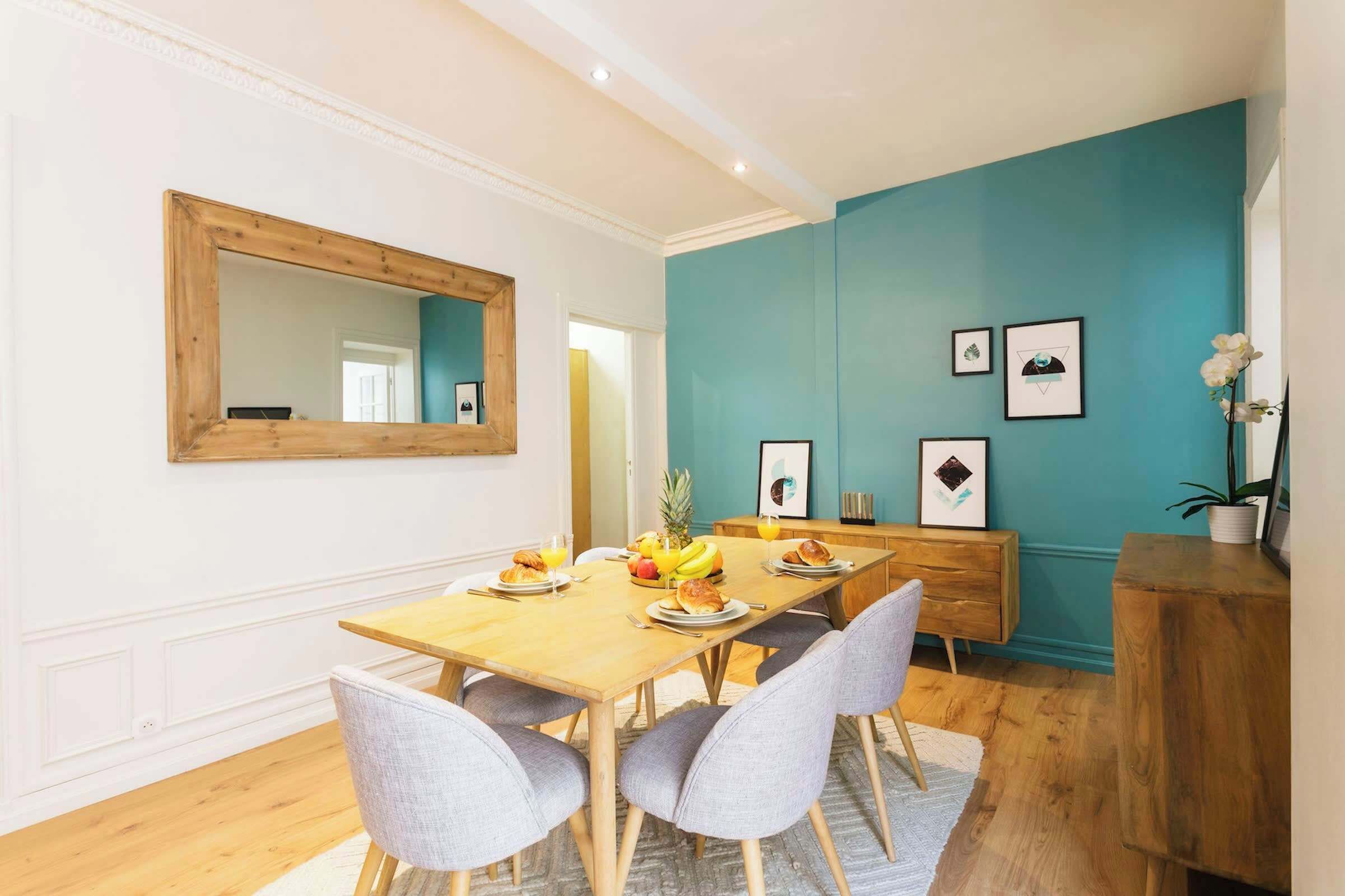 Un coin repas avec une table en bois dressée pour quatre personnes, entourée de chaises grises. Un meuble en bois, surmonté d’un miroir, est adossé au mur de couleur turquoise.