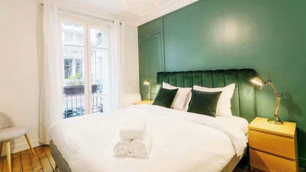 Un lit bien fait, avec du linge de lit blanc et des serviettes pliées, une tête de lit recouverte d’un tissu vert et une lampe de chevet posée sur une table de nuit en bois.