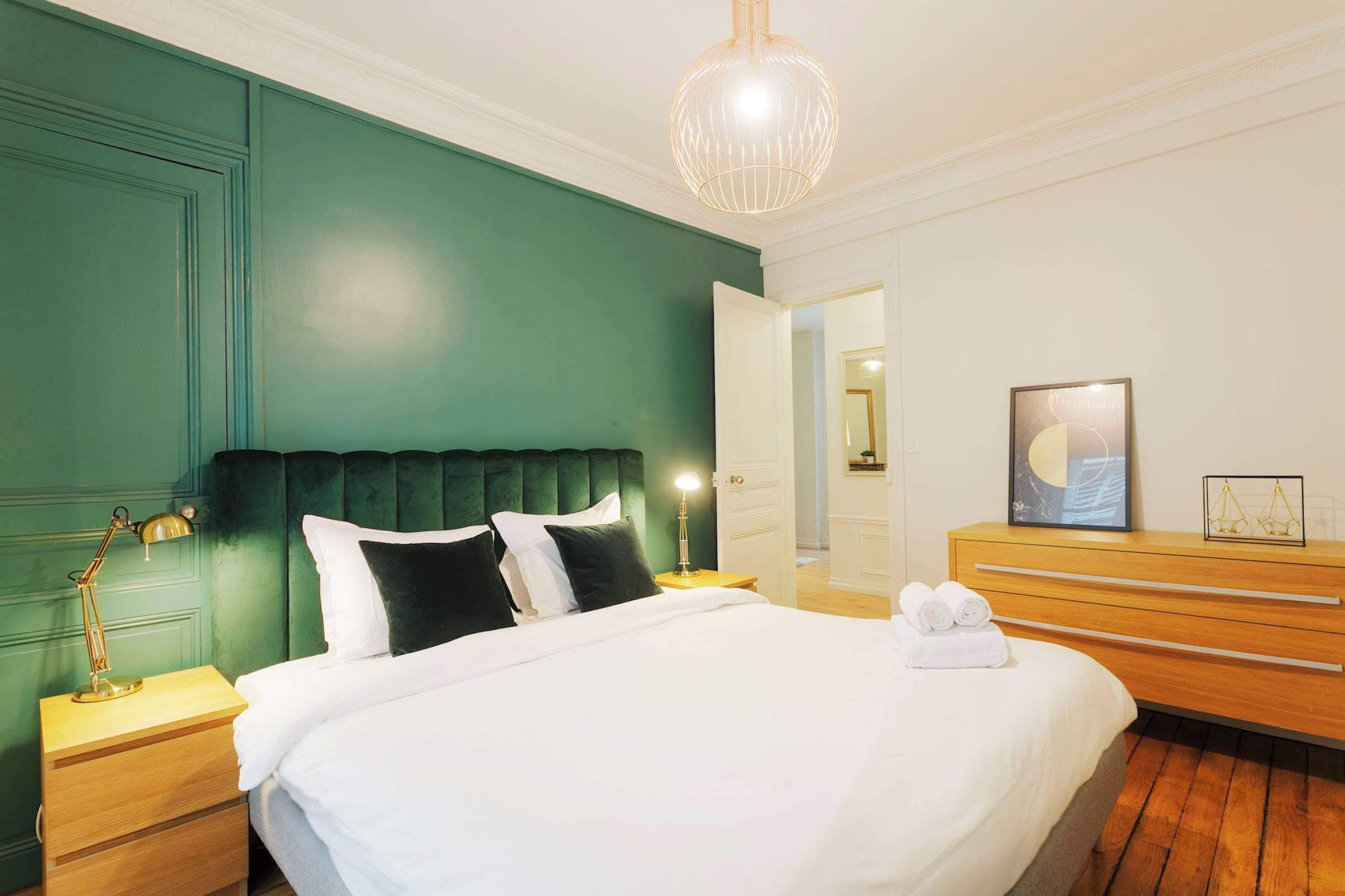 Un lit bien fait, avec du linge de lit blanc, une tête de lit recouverte d’un tissu vert et une table de chevet en bois avec une lampe.