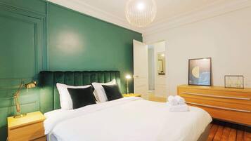 Un lit bien fait, avec du linge de lit blanc, une tête de lit recouverte d’un tissu vert et une table de chevet en bois avec une lampe.