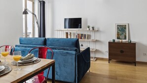 A modern living room with a blue sofa, wooden dining table, and red chairs. A TV is mounted on the wall, and there is a bookshelf with decorative items. - St Germain des Pres - Mabillon 1BR Flat (Paris)