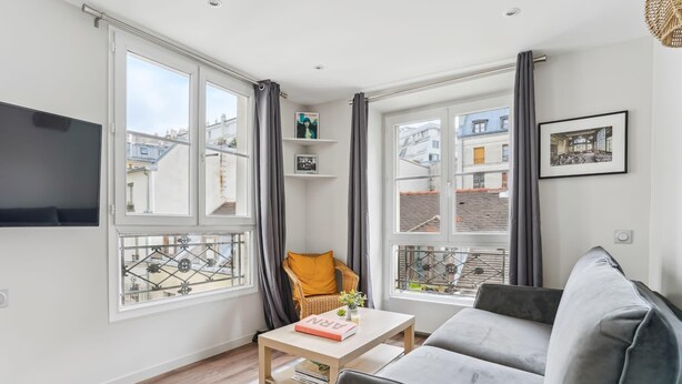 Un salon moderne avec un canapé gris, une table basse en bois sur laquelle repose un livre, un fauteuil en osier et un téléviseur accroché au mur gauche.