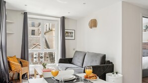 Un salon moderne avec un coin repas, un canapé gris et une vue sur le paysage urbain grâce à de grandes fenêtres.