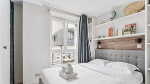 Une chambre moderne avec une tête de lit rembourrée de couleur grise, un lit bien fait avec du linge de lit blanc, une petite étagère avec des livres et des objets décoratifs, et une fenêtre donnant sur un bâtiment à l’extérieur.