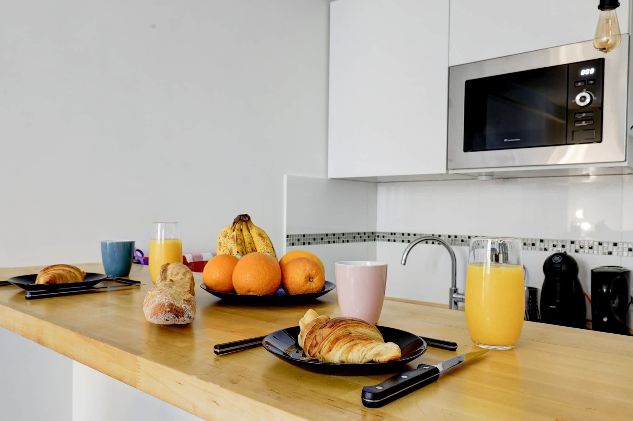 A kitchen counter with breakfast items including croissants, oranges, and juice, a microwave, and a coffee maker.