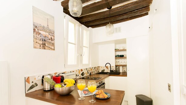 Une cuisine avec un plan de travail en bois, un évier et une fenêtre. On y trouve deux verres de jus d’orange et une assiette avec un croissant posé sur le plan de travail.