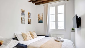 Un lit bien fait, avec un couvre-lit beige et des serviettes pliées, une fenêtre avec des rideaux et des œuvres d’art accrochées au mur.