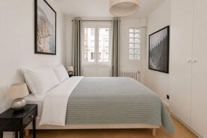A bedroom with a bed, a nightstand, a lamp, a window with curtains, and a wall-mounted picture.