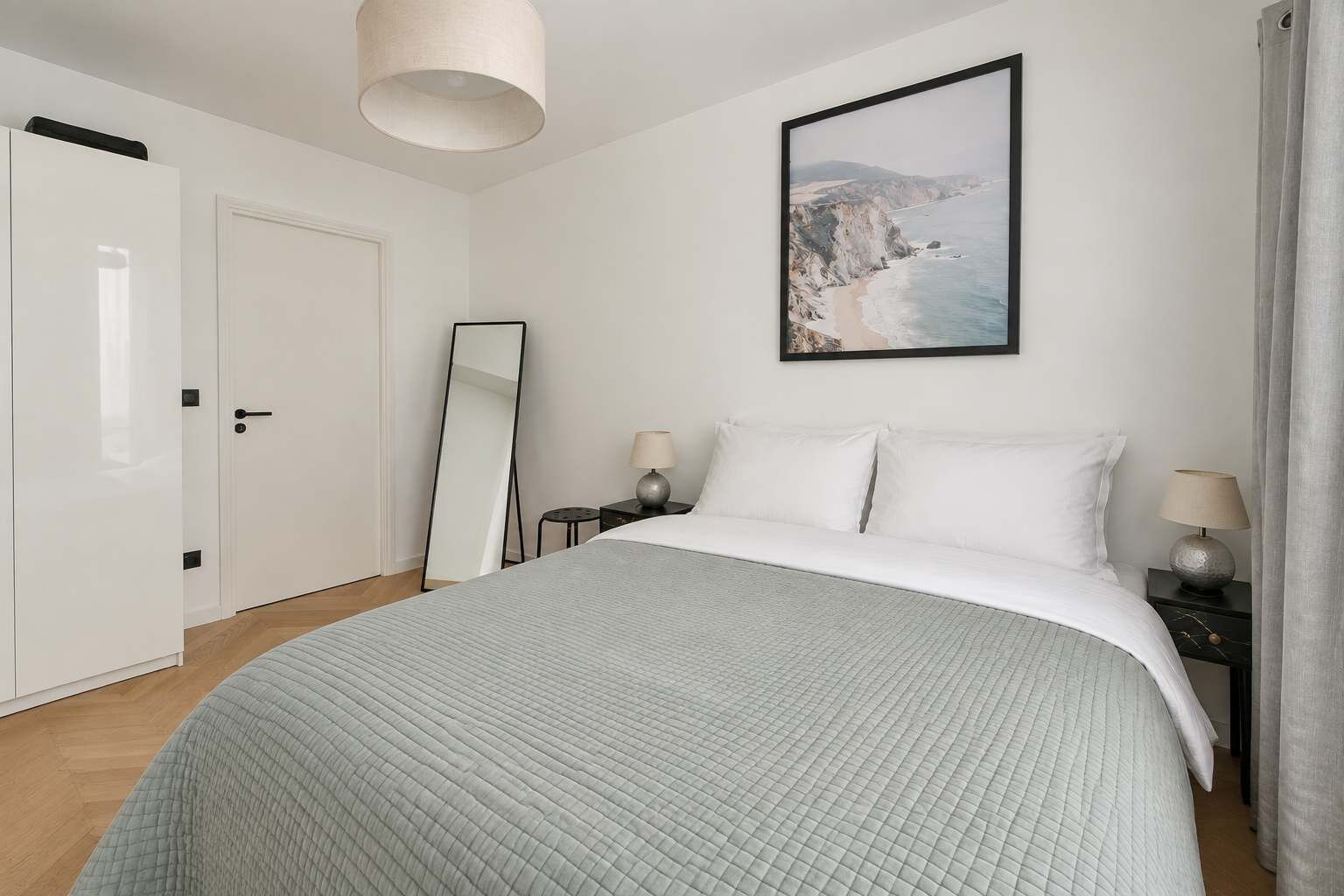 Une chambre à coucher comprenant un lit, une table de chevet, une lampe, un miroir et un tableau représentant un paysage côtier.