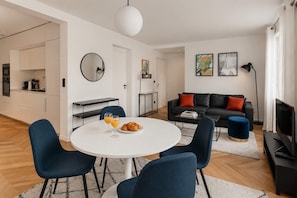 Ein modernes Wohnzimmer mit einem runden Esstisch, blauen Stühlen, einem schwarzen Sofa mit roten Kissen und einem Flachbildfernseher.