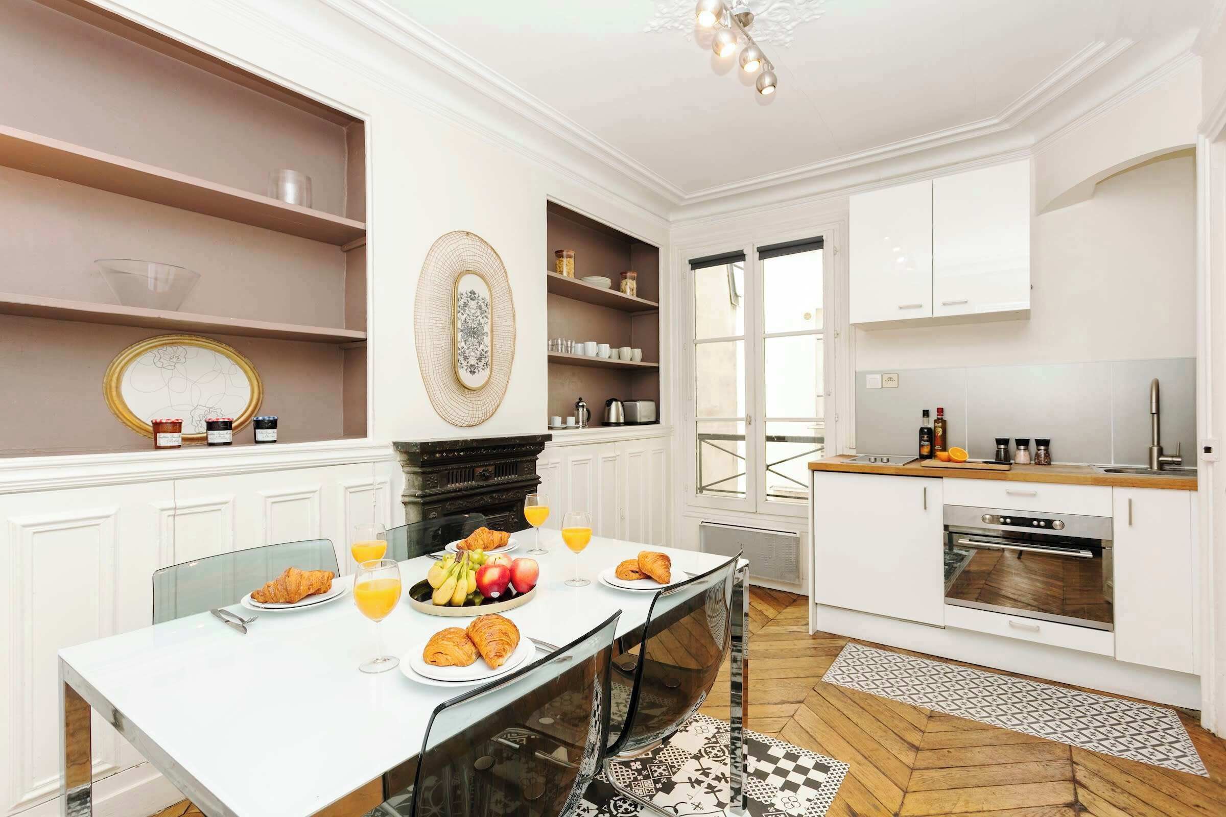 Une cuisine moderne avec des meubles blancs, une table à manger où sont disposés des aliments pour le petit-déjeuner, et une vue sur l’extérieur.