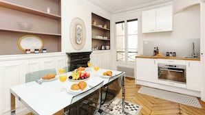 Une cuisine moderne avec des meubles blancs, une table à manger où sont disposés des aliments pour le petit-déjeuner, et une vue sur l’extérieur.