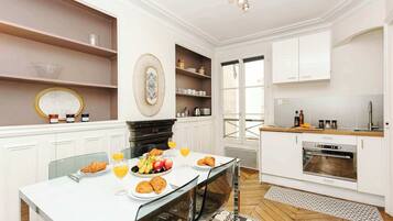 Une cuisine moderne avec des meubles blancs, une table à manger où sont disposés des aliments pour le petit-déjeuner, et une vue sur l’extérieur.