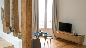 Ein modernes Wohnzimmer mit Holzboden, einem Fernseher auf einem Holzständer, einem kleinen runden Tisch mit Kerzen und Blick ins Freie durch große Fenster.