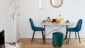 Ein Essbereich mit einem runden Tisch, zwei blauen Stühlen, einem grünen Hocker und einer weißen Wand mit einem hölzernen Kleiderhaken und einem Spiegel.