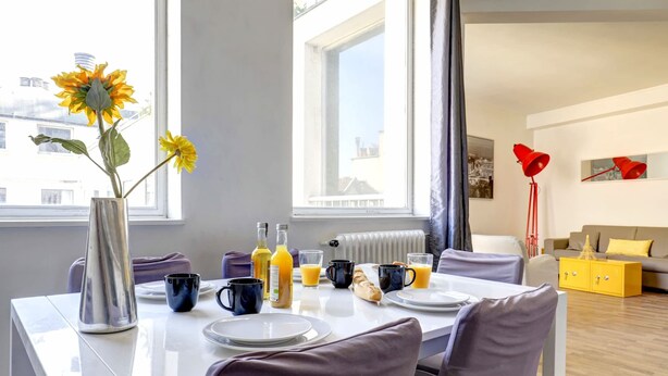 A modern dining area with a white table set for breakfast, a vase with sunflowers, and a window with a view of buildings. - Lovely 1BR Flat by Palais Royal-Le Louvre (Paris)