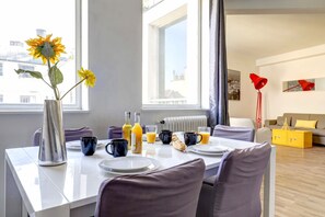 A modern dining area with a white table set for breakfast, a vase with sunflowers, and a window with a view of buildings.