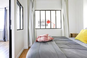 A bedroom with a large window, a bed with grey bedding, a yellow pillow, and a red lamp on the windowsill.