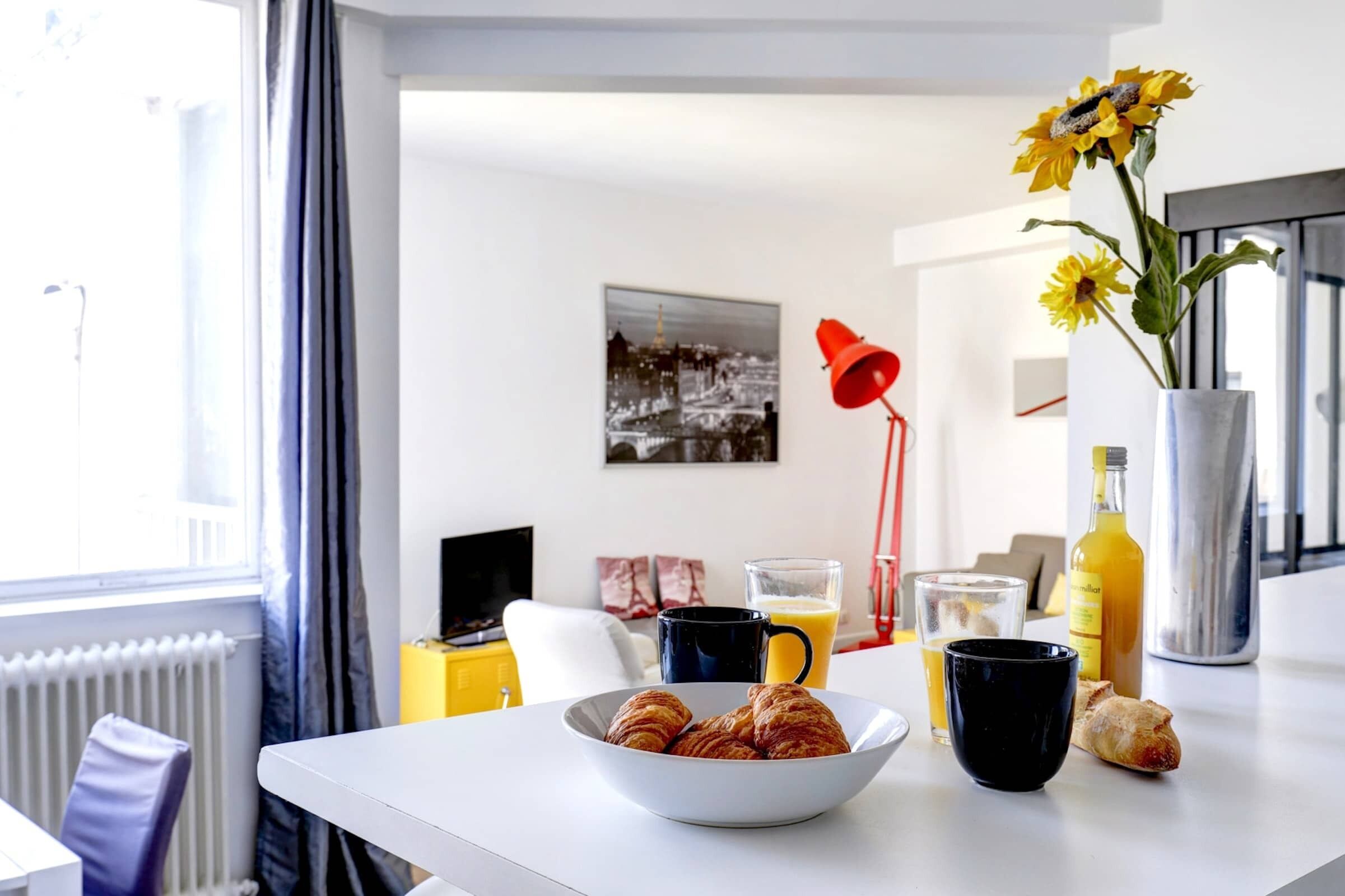 Une table blanche dressée pour le petit-déjeuner, avec des croissants, un bol, une tasse de café, un verre de jus d’orange et une bouteille de jus d’orange.