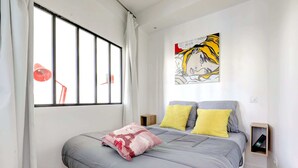A bedroom with a large window, a bed with grey bedding, two pillows, a red lamp, and a wall art piece. - Lovely 1BR Flat by Palais Royal-Le Louvre (Paris)