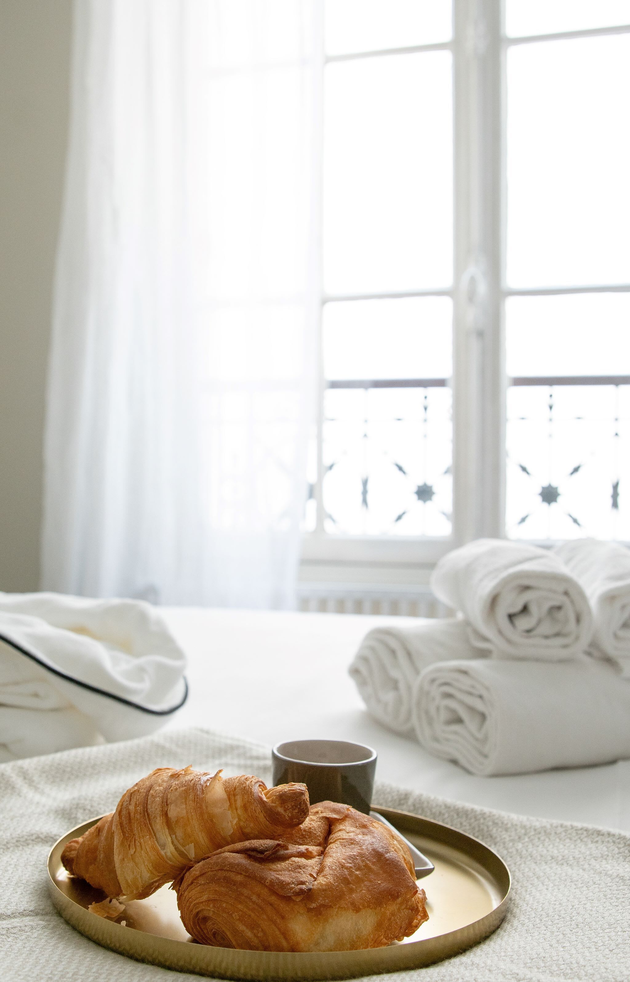 Uma bandeja com croissants e uma xícara sobre uma mesa em um quarto claro, com uma janela e toalhas enroladas ao fundo.
