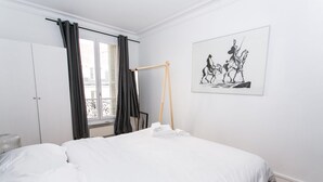 Une chambre à coucher avec un lit, une fenêtre avec des rideaux, un radiateur, un plancher en bois et une œuvre d’art accrochée au mur.