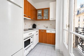 A compact kitchen with white appliances, wooden cabinets, and a view of the street through a window. - Elegant 2BR in St-Germain Near Le Bon Marche (Paris)