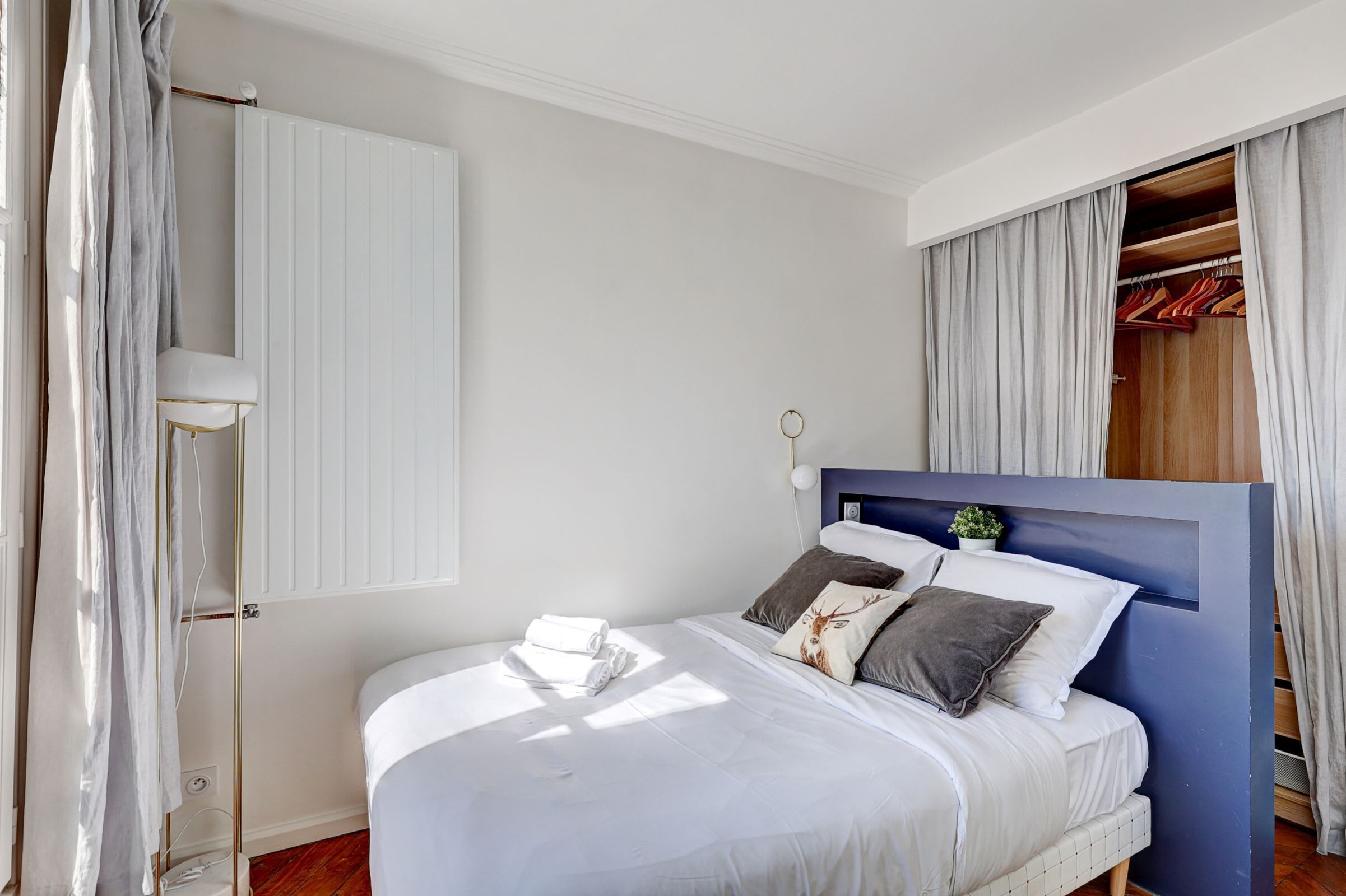 Una cama bien hecha con ropa de cama blanca y almohadas decorativas, una mesita de noche con lámpara y vistas a un armario de madera con ropa colgada.