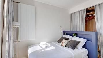 Una cama bien hecha con ropa de cama blanca y almohadas decorativas, una mesita de noche con lámpara y vistas a un armario de madera con ropa colgada.