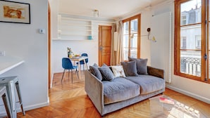 Una sala de estar acogedora con un sofá gris, una mesa de comedor con sillas y un ventanal con vistas a edificios.